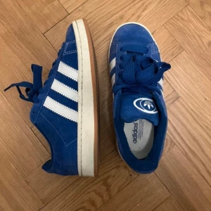 Blå Adidas sneakers - Snygga blå Adidas sneakers med vita ränder och snörning. Skorna har en klassisk design med en gummisula i kontrasterande färg. Perfekta för en avslappnad stil.