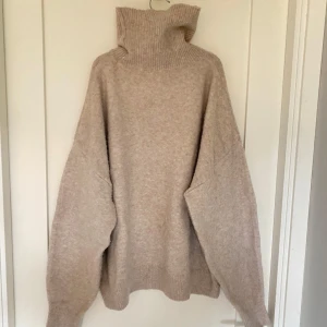 Beige stickad polotröja från H&M - Mysig beige stickad polotröja från H&M med en hög krage. Perfekt för kyliga dagar med sin avslappnade passform och långa ärmar. Tröjan har ribbade detaljer vid krage, ärmslut och nederkant.
