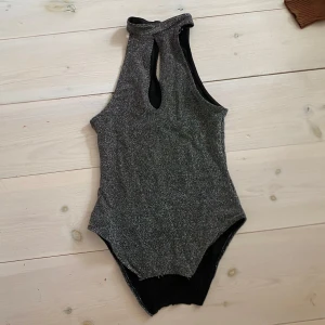 Glittrig body - Snygg glittrig body i silver med hög hals och stilren design. Den har en elegant öppning framtill som ger en extra touch av stil.