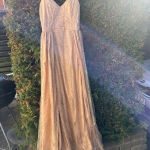 Champagnefärgad långklänning med glittriga detaljer - Elegant champagnefärgad långklänning med v-ringning och glittriga detaljer som ger en lyxig känsla. Klänningen har en vacker fall och är perfekt för speciella tillfällen.