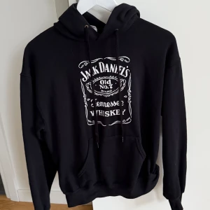 Svart hoodie från Fruit of the Loom - Säljer en svart hoodie från Fruit of the Loom med Jack Daniel's Tennessee Whiskey-tryck på framsidan. Tröjan har en klassisk känguruficka och justerbar huva med dragsko. Perfekt för en avslappnad stil.