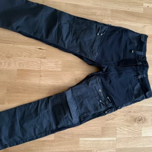 Svarta byxor från Haunter - Säljer ett par svarta byxor från Haunter med flera praktiska fickor och justerbara remmar. Perfekta för utomhusaktiviteter med robust design och dragkedjor. Passar bra för den som söker funktionella och stilrena byxor. Något liten i storleken, mer som strl 40.