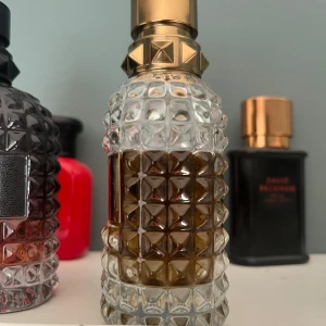 Lyxig parfymflaska med nitar - Elegant parfymflaska med ett unikt mönster av nitar och en gyllene kork. Flaskan har en transparent design som visar den bärnstensfärgade vätskan inuti. Perfekt för den som vill ha en stilfull och iögonfallande parfym på hyllan.