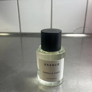 Vanilla Sass Eau de Parfum från ESSNCE - Upptäck Vanilla Sass Eau de Parfum från ESSNCE, en doft med en söt och kryddig vaniljton. Flaskan är stilren och genomskinlig med en svart kork, perfekt för den som älskar en klassisk och elegant doftupplevelse.