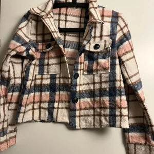 Rutig croppad jacka från Ullared - Säljer en snygg rutig jacka från Ullared i vit, blå och rosa. Jackan är croppad med knappar framtill och har två bröstfickor med knappar. Perfekt för en trendig look!