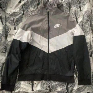Grå och svart vindjacka från Nike - Säljer en snygg grå och svart vindjacka från Nike med vit detalj över bröstet. Jackan har dragkedja framtill och två fickor med dragkedjor. Perfekt för blåsiga dagar och har en huva för extra skydd.
