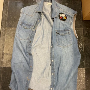 Jeansväst med punkmärke - Säljer en cool jeansväst med ett stort punkmärke på bröstet. Västen har en klassisk krage och knappar framtill. Perfekt för dig som vill ha en edgy stil.