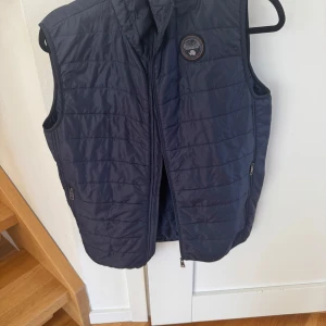 Mörkblå quiltad väst från Napapijri - Snygg mörkblå väst från Napapijri med dragkedja och rund logga på bröstet. Perfekt för lager-på-lager under kyliga dagar. Men även perfekt för ett tunt lager över en tröja på sommaren. Västen har en stilren design med praktiska fickor. Storlek:162. Nypris:1395kr. Säljer för 599kr. Bra skick. Pris kan diskuteras