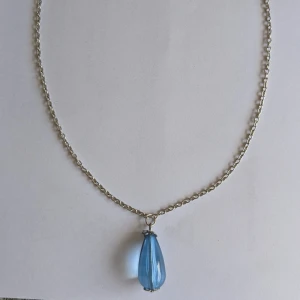 Halsband med blå droppe - Elegant halsband med en vacker blå droppe som hänge. Kedjan är tunn och smidig, vilket gör den perfekt för att framhäva det blå hänget. Passar utmärkt för att ge en färgklick till din outfit.