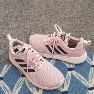 Ljusrosa Adidas sneakers med mesh - Snygga ljusrosa Adidas sneakers med svart logga och klassiska tre ränder på sidorna. Ovandel i ventilerande mesh, vit platt sula och snörning. Perfekta för dig som gillar sportig och fräsch stil. Cloudfoam-dämpning för extra komfort.