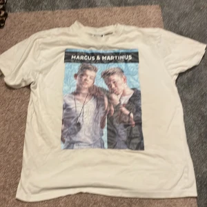 Vit Marcus & Martinus t-shirt MMSTORE - Vit t-shirt med tryck av Marcus & Martinus på bröstet och svart text. Klassisk passform med rund hals och korta ärmar. Perfekt för fans som vill visa sin stil. Materialet känns mjukt och skönt mot huden.  har några fläckar på ryggen 