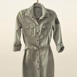 Grön skjortklänning med bälte och fickor - Snygg militärgrön skjortklänning från ESPRIT by EDC med knappar framtill, två bröstfickor och ett bälte i midjan. Klänningen har långa ärmar och dekorativa detaljer som nitar och skraphålslappar. Perfekt för en cool och avslappnad stil.