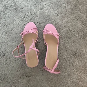 Rosa sandaler med rosett och klack - Säljer ett par ljusrosa sandaler med liten klack och öppen tå. Skorna har en söt rosettdetalj framtill och smala remmar med spänne runt vristen. Perfekta för vår och sommar! Stolek 37, helt oanvända. 