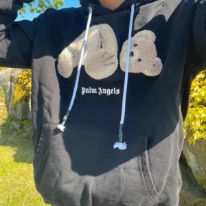 Svart hoodie från Palm Angels med nallemotiv - Svart hoodie från Palm Angels med stort broderat nallemotiv på bröstet och vit logotyp under. Tröjan har huva med dragsko och en stor magficka framtill. Perfekt för dig som gillar streetwear och vill sticka ut.