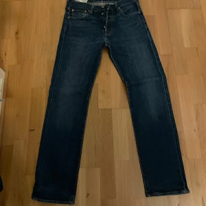 Mörkblå raka jeans från Levi's - Klassiska mörkblå jeans från Levi's med raka ben och femficksdesign. Jeansen har orange sömmar, knappgylf och den ikoniska läderpatchen bak i midjan. Passar perfekt till vardags och har en tidlös look. Stl 30/32
