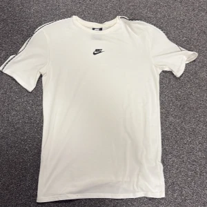 Vit Nike t-shirt med logga, S - Vit t-shirt från Nike i bomull med klassisk svart Nike-logga på bröstet och svarta detaljer längs axlarna. Modellen är standard fit med rund hals och korta ärmar. 