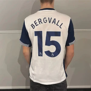 Tottenham Hotspur Bergvall  - Säljer en Tottenham Hotspur fotbollströja med namn och nummer Bergvall 15. Tröjan är vit med mörkblå ärmar, röda AIA-trycket på bröstet och Premier League-logga på ärmen. Klassisk Nike-design och klubbmärke på framsidan.