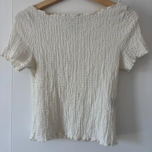 Vit ribbad topp - Säljer en söt vit topp med ribbad struktur och korta ärmar. Toppen har en lätt croppad passform och vågig kant vid halsringning och nederkant. Perfekt till sommaren och enkel att matcha med olika outfits. Den är i storlek M men skulle påstå att den snarare är S i passform.