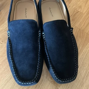 Marinblå mocka loafers från Gant - Säljer ett par stilrena marinblå loafers från Gant i mjuk mocka. Skorna har kontrasterande vit söm runtom och klassisk design med rund tå. Perfekta för en dressad look och passar till många tillfällen. Ordinarie pris 1500 kr, endast använda en gång eftersom det var en för stor storlek. Pris går att diskutera. 
