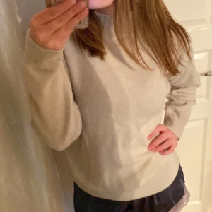 Beige stickad tröja med hög krage - Mysig beige stickad tröja med hög ribbad krage och långa ärmar. Tröjan har en relaxed passform och breda ribbade muddar vid ärmslut och nederkant. Perfekt för lager-på-lager under kyliga dagar.