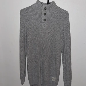 Grå stickad tröja från Division Rads - Säljer en grå stickad tröja från Division Rads med hög krage och tre knappar framtill. Tröjan har lång ärm och en liten patch nertill. Perfekt för kyligare dagar och enkel att matcha med jeans eller chinos.