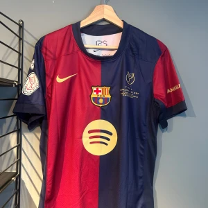 FC Barcelona Lamine Yamal matchtröja - Snygg FC Barcelona matchtröja med Lamine Yamal och nummer 19 på ryggen. Tröjan är kortärmad och har klassiska blå och röda fält, guldiga detaljer och tryck. Officiella klubbmärket, Nike-logga och Spotify-sponsor framtill. Tillverkad i lätt och ventilerande material. Helt ny med tagg en replika 