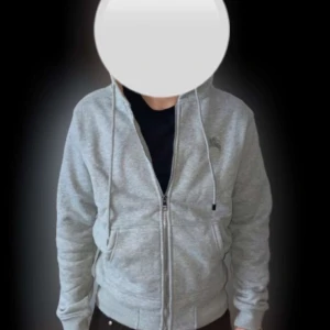 Grå hoodie från Burberry med dragkedja - Säljer en grå hoodie från Burberry med dragkedja och klassiskt rutigt foder i huvan. Tröjan har fickor framtill och justerbar huva med snören. Perfekt för en stilren och avslappnad look. (Har aldrig använts)