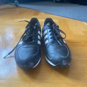 Svarta Adidas LA Trainer sneakers - Säljer ett par svarta Adidas LA Trainer sneakers med vita ränder och röda detaljer på sulan. Skorna har snörning, rund tå och klassisk retrostil. Perfekta för dig som gillar sportig och stilren look. Lite dammiga då de legat nerpackade ett tag. 