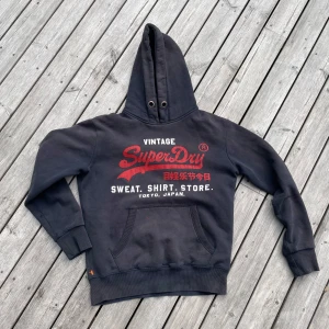 Mörkgrå hoodie från Superdry - Säljer en riktigt snygg mörkgrå hoodie från Superdry med röd och vit logga på bröstet. Tröjan har känguruficka framtill, huva med öljetter och ribbade muddar. Den har ett litet hål vid armbågen. Skriv gärna vid funderingar 