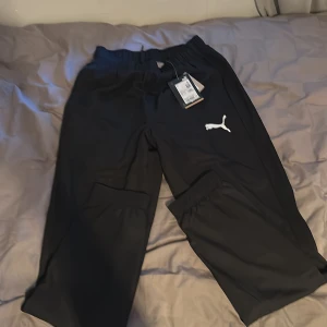 Svarta träningsbyxor från Puma - Svarta mjukisbyxor från Puma med elastisk midja och vit logga på benet. Byxorna är tillverkade i polyester och har en sportig look med meshfoder på insidan. Perfekta för träning eller chill.