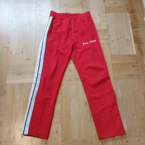 Röda Palm Angels mjukisbyxor - Röda mjukisbyxor från Palm Angels med vit sidoremsa och svart rand. Byxorna har elastisk midja och raka ben, samt logga broderad på vänster sida. Perfekta för en sportig och trendig look. Har en liten svart prick på höger knä syns nog inte ens i bild 