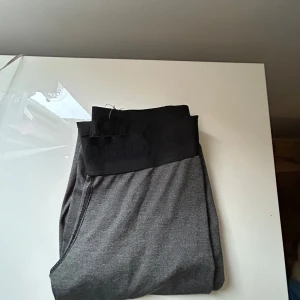 Grå Adidas leggings med logga - Mörkgrå leggings från Adidas med bred svart resår i midjan och stor vit Adidas-logga längs benet. Stretchigt material som sitter tight och passar perfekt till träning eller chill. Snygg och sportig look med diskreta sömmar.