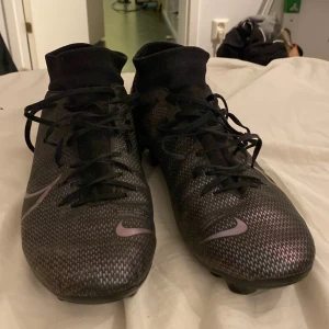 Nike Mercurial academy svarta fotbollsskor - Svarta Nike Mercurial fotbollsskor med silvriga detaljer och snörning. Skorna har en strumpliknande hög krage och mönstrad ovandel i syntetmaterial. Perfekta för dig som vill ha grymt grepp på planen och snygg stil.