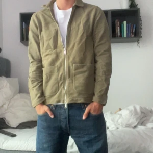 Beige overshirt från Riley - Säljer en stilren beige overshirt från Riley med dragkedja framtill och tre stora fickor. Jackan är i bomull och har en avslappnad passform, perfekt att slänga på över en t-shirt. Enkel och clean look som funkar till det mesta.