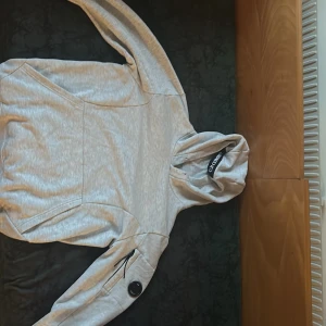 Grå hoodie C.P. Company - Säljer en stilren ljusgrå hoodie från C.P. Company med klassisk huva och logotyp på ärmen. Tröjan har lång ärm, ribbade muddar och en praktisk ficka med dragkedja på ärmen. Perfekt för dig som gillar streetwear och snygga detaljer.