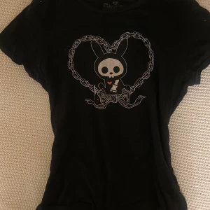 Svart t-shirt med kanintryck - Svart t-shirt med korta ärmar och tryck av en söt kanin med dödskalleansikte inramad av ett hjärta i kedjor. T-shirten har en smal passform och är perfekt för dig som gillar alternativ stil. Materialet känns mjukt och bekvämt. Köpt i USA på Hot Topic och använd ett par gånger. 