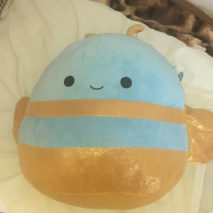 Queen Bee - Möt Queen Bee från Squishmallows! En supermjuk och gosig gosedjurskudde i blått och guld med ett gulligt leende. Perfekt för att mysa med eller som en cool inredningsdetalj i ditt rum. Passar dig som älskar söta och trendiga samlarprylar.