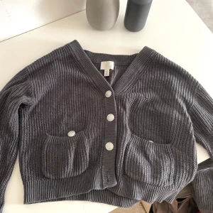 Grå kofta med fickor - Grå stickad kofta från H&M med croppad passform, stora framfickor och silvriga knappar. Långärmad och ribbad i både ärmslut och nederkant. Enkel att styla till jeans eller kjol. Nedersta knapp trasig.