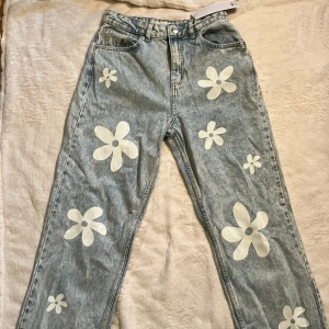 Blommiga jeans - Unika ljusblå jeans från FB Sister i storlek S med stora vita blommor tryckta över hela byxorna. Modellen är rak och har klassisk femficksdesign, bälteshällor och knapp med blomdetalj. Perfekta för dig som vill sticka ut med en lekfull och trendig look.
