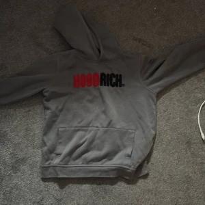 Grå hoodie från Hoodrich - Säljer en grå hoodie från Hoodrich med tryck i rött och svart på bröstet. Tröjan har huva och en stor magficka framtill. Passformen är normal och materialet känns mjukt och bekvämt, perfekt för vardagsbruk.