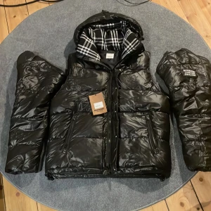 Burberry pufferjacka  - en svart pufferjacka från Burberry med avtagbara ärmar och klassiskt rutigt foder. Jackan har en dragkedja framtill och en stilren logotyp på bröstet. Perfekt för kyliga dagar den är omsyd så den är lite tajtare men fortfarande en stor jacka. (KÖP / BYT) kommer ta ner anonnsen snart så skyna på!