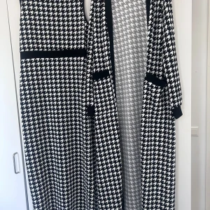 Svartvit ärmlös klänning med matchande lång kofta - Säljer en svartvit ärmlös klänning med klassiskt hundtandsmönster och svart midjeband. Till klänningen hör en lång kofta i samma mönster med svarta detaljer vid kanter och fickor. Perfekt för dig som gillar tidlös stil.