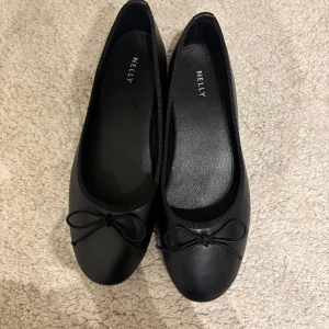 Svarta ballerinaskor med rosett från Nelly - Klassiska svarta ballerinaskor från Nelly med rund tå och en söt rosettdetalj framtill. Skorna har platt sula och är tillverkade i syntetmaterial, perfekta för en stilren och enkel look. Säljes pga för stora. 