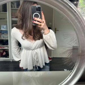 Vit blus - Säljer en söt vit omlottopp med knyt i midjan och fina spetsdetaljer vid urringningen. Toppen har lång ärm och är tillverkad i ett mjukt, ribbat material som ger en skön och avslappnad känsla.