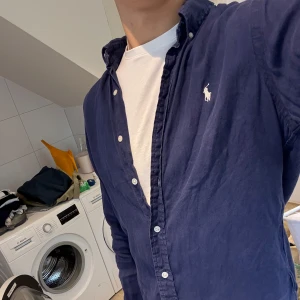 Blå Polo Ralph Lauren Linneskjorta - Snygg blå skjorta från Ralph Lauren i 100% linne, slim fit-modell, storlek M. Klassisk button-down krage, vita knappar och ikoniska broderade loggan på bröstet. Perfekt för sommarn!