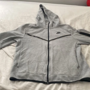 Grå Nike hoodiejacka med dragkedja - Snygg grå hoodiejacka från Nike med hel dragkedja, svart Nike-logga på bröstet och svarta detaljer längs bröstet. Jackan har huva och en praktisk ficka med dragkedja på ärmen. Perfekt för chill eller träning.