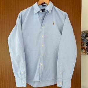 Ljusblå skjorta från Ralph Lauren - Klassisk ljusblå skjorta från Ralph Lauren med button-down krage och broderad logga på bröstet. Skjortan har långa ärmar, normal passform och stängs med vita knappar framtill. Perfekt för en clean och stilren look.