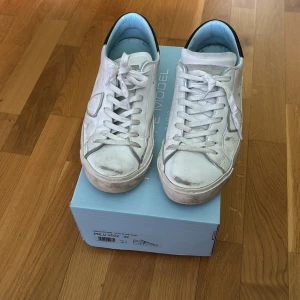 Vita Philippe Model sneakers, strl 42 - Snygga vita sneakers från Philippe Model med svarta detaljer på hälen och ljusblå insida. Klassisk låg modell med snörning och rund tå. Tillverkade i skinn med diskret logga på sidan. Perfekta för en clean och stilren look.