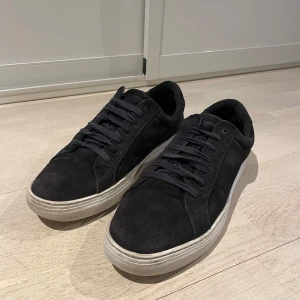 Svarta mockasneakers från Vagabond - Stilrena svarta sneakers från Vagabond i mjuk mocka med klassisk vit sula. Skorna har rund tå, svarta skosnören och en enkel, clean design som passar till det mesta. Perfekta för dig som gillar minimalistisk stil och vill ha ett par bekväma vardagssneakers.