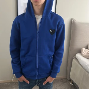 Blå hoodie Comme des Garçons Play - Blå hoodie med dragkedja från Comme des Garçons Play. Ikoniskt svart hjärta med ögon på bröstet och stort hjärta på ryggen. Huva, känguruficka och ribbade muddar. Perfekt för dig som gillar streetwear och vill sticka ut.